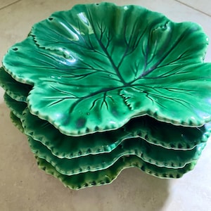 May include: A stack of four green ceramic leaf-shaped plates. Each plate has a detailed vein pattern and a scalloped edge. The plates are a vibrant shade of green, suggesting a natural, botanical theme.