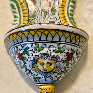 May include: Hand-painted ceramic wall pocket with a detailed floral and cherub design. The piece features a white base with yellow, green, and blue accents. The cherub has yellow skin and blue wings. The wall pocket is a decorative item.