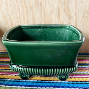 May include: A dark green ceramic planter with a square shape and a decorative, fluted base with three scroll feet.
