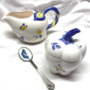 May include: A white ceramic creamer, sugar bowl, and spoon set. The creamer and sugar bowl are decorated with blue and yellow floral and butterfly designs. The spoon has a butterfly image.
