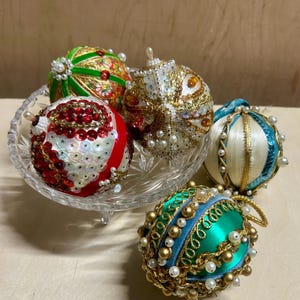 May include: Five spherical ornaments decorated with beads, sequins, and metallic trim. One is green and red, one is white and gold, one is red and white, one is cream and teal, and one is teal and gold. They are arranged in a clear glass bowl.