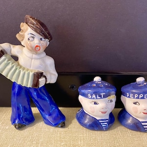 May include: A vintage ceramic salt and pepper shaker set featuring a musician playing an accordion and two sailor-themed shakers. The musician is wearing a white shirt, blue pants, and a brown beret. The sailor shakers are blue and white.
