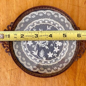 May include: A round, antique-style frame with a delicate white lace design inside. The frame is made of metal with a brown finish and has two ornate handles. The frame measures 6 inches in diameter.