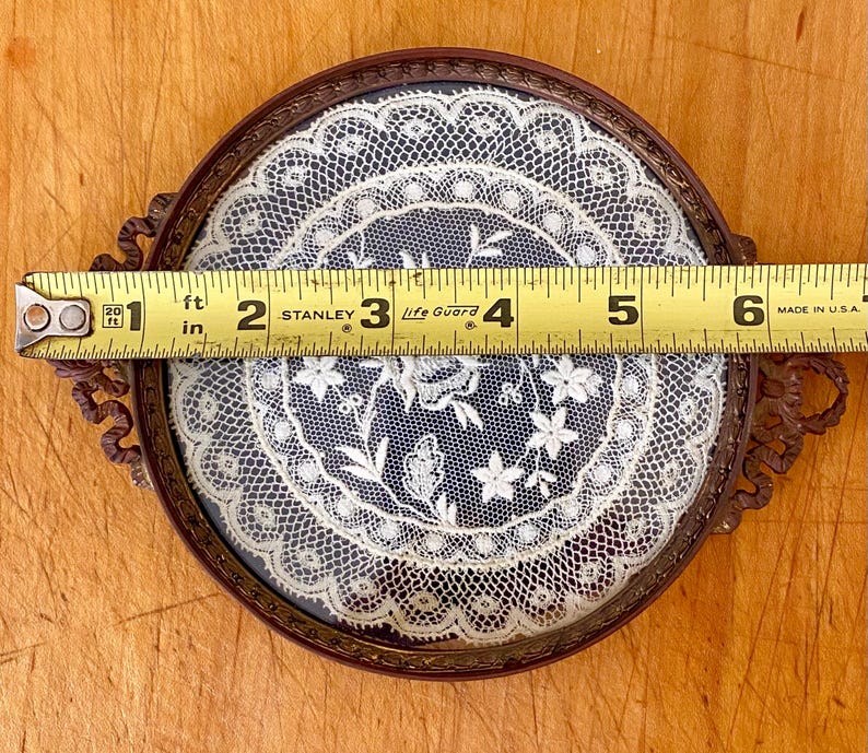 May include: A round, antique-style frame with a delicate white lace design inside. The frame is made of metal with a brown finish and has two ornate handles. The frame measures 6 inches in diameter.