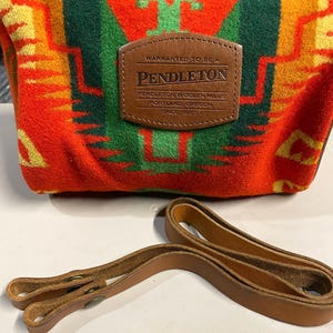 May include: A red, green, and yellow Pendleton blanket with a brown leather strap. The blanket has a traditional Native American pattern and the strap has two metal buckles. The blanket has a leather tag that reads "Warranted to be a Pendleton, Pendleton Woolen Mills, Portland, Oregon, Since 1883."