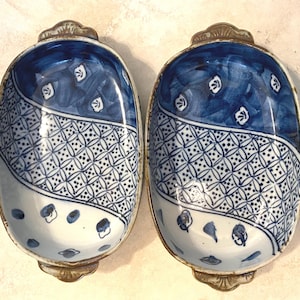 May include: Two blue and white ceramic serving dishes with a decorative pattern. Each dish has a silver-colored metal handle and rim. The dishes are oval-shaped and feature a split design with a geometric pattern.
