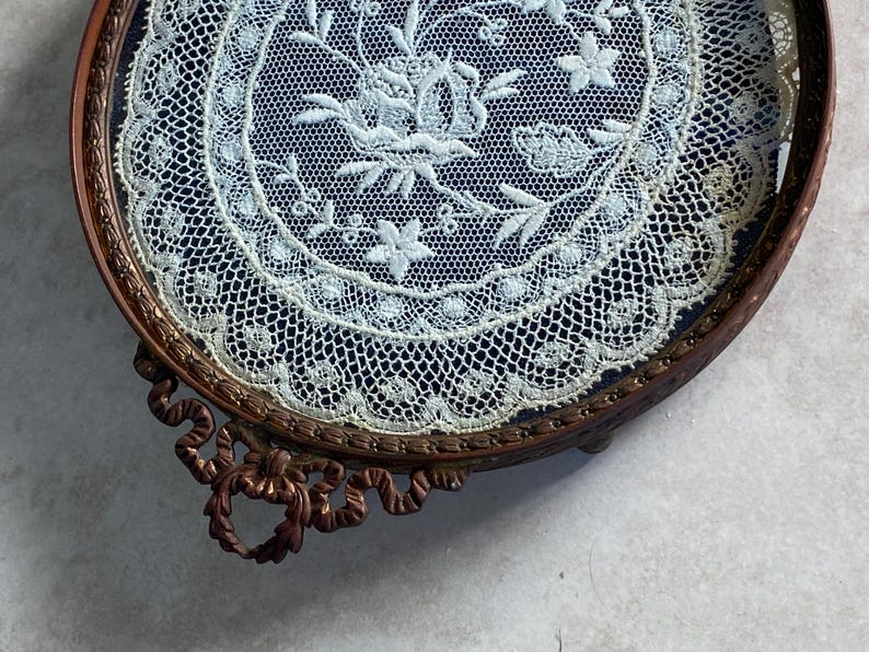 May include: A round, antique, brass tray with a delicate lace design inside. The tray has a decorative, ornate, scroll-like base.