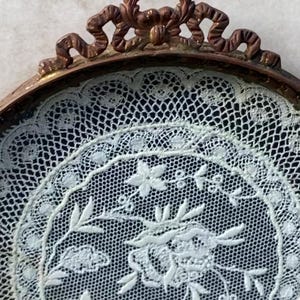 May include: A white lace doily with a floral design is displayed in a round, ornate, copper-colored frame.