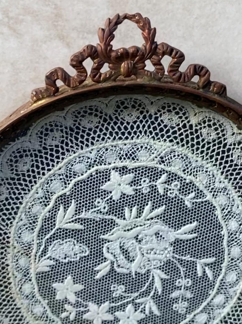 May include: A white lace doily with a floral design is displayed in a round, ornate, copper-colored frame.