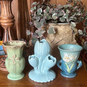 May include: Three ceramic vases arranged in a row. The left vase is light green with a flared rim and floral relief. The center vase is light blue with a three-part tulip shape. The right vase is blue-green with a white iris flower relief.