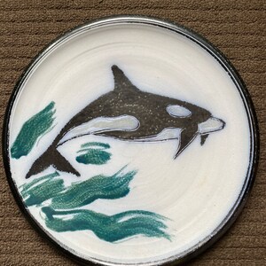 May include: A white ceramic plate with a black and white orca whale design. The orca is swimming through green seaweed. The plate has a black rim.