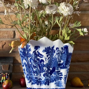 May include: A blue and white ceramic vase with a scalloped rim and a floral design. The vase features a scene of people and a bridge in a landscape. The vase is filled with white flowers.