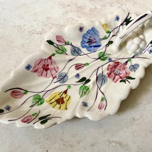 May include: A white ceramic leaf-shaped serving dish with a handle. The dish is decorated with a floral pattern in shades of pink, blue, yellow, and green.