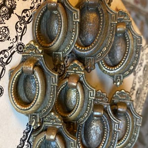 May include: A collection of seven antique-style brass drawer pulls featuring a shield-shaped backplate with a textured oval center and a swinging circular ring handle. Each piece displays a weathered metallic finish with intricate decorative detailing.