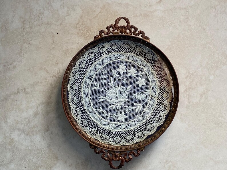 May include: A vintage, round, metal tray with a delicate lace design inside. The tray has a dark brown finish and ornate scrollwork around the edge.