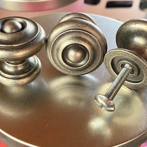 May include: Close-up of four antique bronze cabinet knobs. The knobs have a round, decorative design with a screw and washer visible on one. The knobs are arranged on a silver surface.
