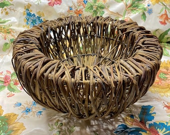 May include: A round, woven basket made of brown twigs. The basket has a wide, open design with a circular shape. The twigs are tightly woven together, creating a textured surface. The basket is suitable for decorative or storage purposes.