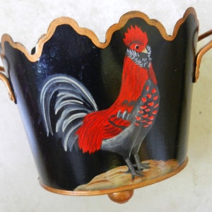 May include: A black metal planter with a scalloped rim and two copper handles. The planter features a hand-painted rooster with red, black, and white feathers. The planter has a copper base with a single foot.