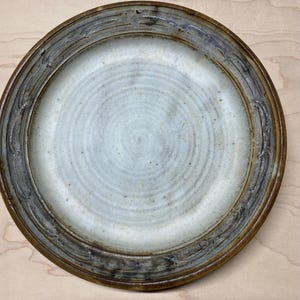 May include: A white ceramic plate with a brown rim and a decorative design. The plate has a subtle swirl pattern in the center.