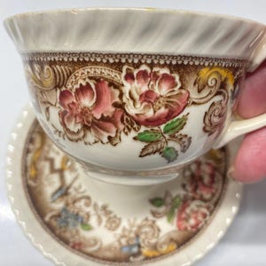 May include: A vintage teacup and saucer set with a floral pattern in shades of brown, pink, and green. The cup and saucer have a white background with a scalloped edge.