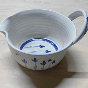 May include: A white ceramic mixing bowl with a pouring spout and a single handle. The interior base features a blue circular band with painted blue floral sprigs. The exterior is decorated with a repeating pattern of small blue painted flowers.
