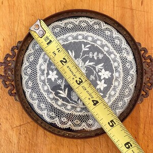 May include: A round, brass-framed tray with a delicate white lace doily inside. The tray has two ornate handles and measures approximately 3.5 inches in diameter.