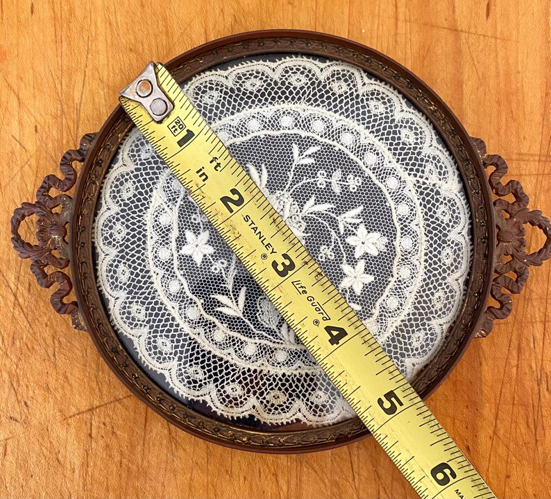May include: A round, brass-framed tray with a delicate white lace doily inside. The tray has two ornate handles and measures approximately 3.5 inches in diameter.