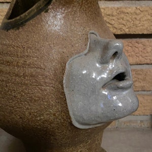 May include: A ceramic sculpture of a face with a brown and gray glaze. The face is partially obscured by a brown, textured, ceramic form with two horns.