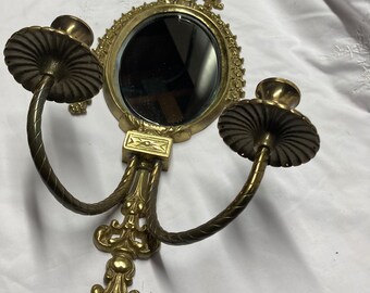 May include: Gold-toned wall sconce with two candle holders and a small oval mirror in the center. The sconce has a decorative design with intricate details.