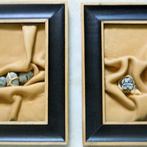 May include: Two framed pieces of art with a tan leather background and gray stones. The stones are arranged in a way that suggests they are being held by the leather.
