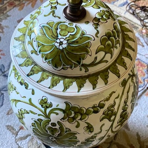 May include: A ceramic table lamp base with a rounded body and a matching lid, featuring a white glazed finish decorated with hand-painted green floral patterns and vine motifs. A brass-colored metal rod extends from the center of the lid.