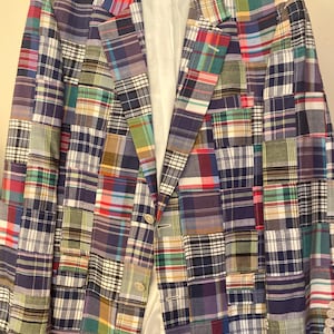 May include: A blazer constructed from a patchwork of various plaid fabrics in colors including red, blue, green, yellow, and white. The garment features a notched lapel, a two-button front closure, long sleeves with four buttons at the cuffs, and a white lining.
