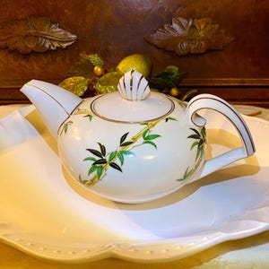 May include: A white porcelain teapot with a bamboo design in green and gold. The teapot has a curved handle and spout, and a shell-shaped lid finial. It sits on a white, scalloped plate.