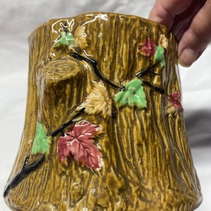 May include: A ceramic planter shaped like a tree stump with brown, green, and red leaves. The planter has a textured surface to resemble bark.