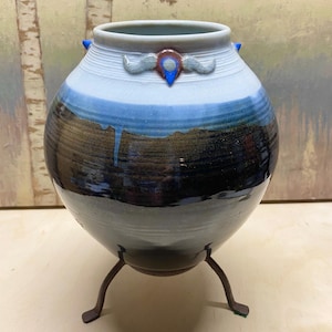 May include: A round ceramic vase with a wide opening, featuring a gradient glaze that transitions from white at the top to deep blue and dark brown at the base. The vase is supported by a three-legged metal stand and has a small decorative emblem.