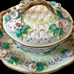 May include: A white porcelain butter dish with a lid and two handles. The dish is decorated with flowers and leaves in shades of green, pink, yellow, and blue. The lid has a brown handle.