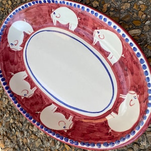 May include: An oval ceramic serving platter with a red and white glaze. The platter is decorated with a border of blue dots and a repeating pattern of white pigs.