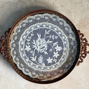 May include: A vintage brass tray with a glass insert featuring a delicate white lace doily with a floral design.