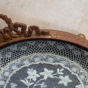 May include: A round, ornate, gold-toned frame with a delicate white lace design inside. The frame has a decorative, scroll-like design around the edge.