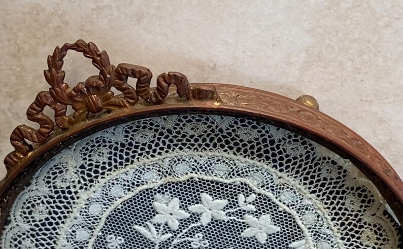May include: A round, ornate, gold-toned frame with a delicate white lace design inside. The frame has a decorative, scroll-like design around the edge.