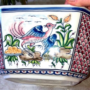 May include: A white ceramic planter with a hexagonal shape featuring a hand-painted scene of a bird with blue and pink wings, green foliage, and brown rocks. The side panels have a repeating pink and white scale pattern with blue borders.