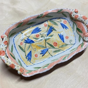 May include: A rectangular ceramic tray with scalloped edges and two handles. The interior features a yellow background with hand-painted blue bell-shaped flowers, green leaves, and orange dots. The rim is accented with peach and black polka dots.