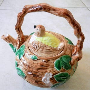 May include: A ceramic teapot shaped like a bird's nest with a bird on the lid. The teapot is brown with green leaves and pink flowers. The bird is yellow with brown wings.