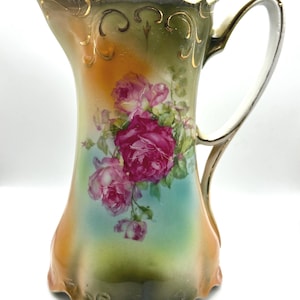 Puede incluir: Una jarra de porcelana vintage con un diseño floral. La jarra está decorada con rosas rosas y rojas, hojas verdes y detalles dorados. La jarra tiene una base festoneada y un asa.
