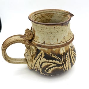 Pode incluir: Caneca de cerâmica artesanal com um design único. A caneca apresenta uma metade superior bege clara e uma metade inferior com padrões abstratos marrons escuros. Possui uma alça grande com redemoinhos decorativos e um pequeno bico.