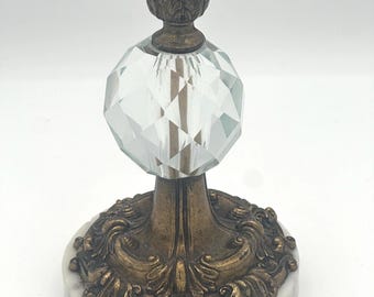 May include: A vintage candle holder with a crystal ball and a gold metal base with ornate details. The base is set on a white marble platform.