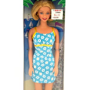 Puede incluir: Una muñeca Barbie en una caja. La muñeca tiene el pelo rubio y lleva un vestido azul con estampados de margaritas blancas y tirantes amarillos. La caja tiene texto que dice "Edición Especial" en varios idiomas.