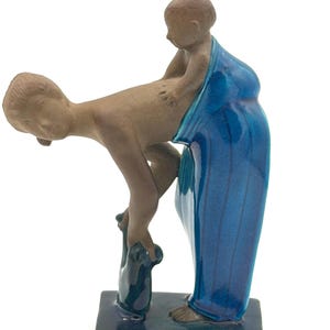 Puede incluir: Una escultura de cerámica que representa una figura sosteniendo a un niño, envuelta en una tela azul. Las figuras son de color marrón, colocadas sobre una base rectangular azul oscuro. La escultura tiene un acabado brillante.