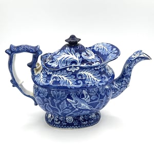 Pode incluir: Uma chaleira azul e branca com um design floral e um motivo de pássaro. A chaleira tem um bico único e curvo e uma tampa com um botão.