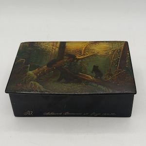 May include: A rectangular black box with a forest scene on the lid. The lid features a painting of bears in a forest with trees and a fallen log. The box has text on the side.